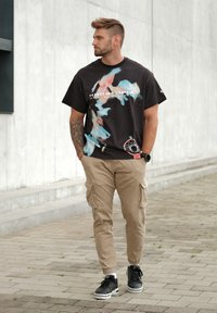 Czarna koszulka z kolorowym abstrakcyjnym wzorem i napisem "HOOPTOPIA ŻYCZYMY, ABYŚ BYŁ TU;" w połączeniu z beżowymi spodniami cargo i czarnymi sneakersami.