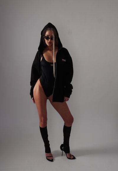 Sudadera negra con cierre, sobre un traje de baño negro de una sola pieza, calcetas negras hasta la rodilla y sandalias negras de tacón alto. Diseño elegante y ajustado.