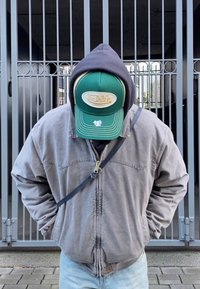 Giacca di denim grigia con zip, indossata sopra un hoodie scuro. Cappello verde con logo bianco, in piedi davanti a un cancello di metallo.