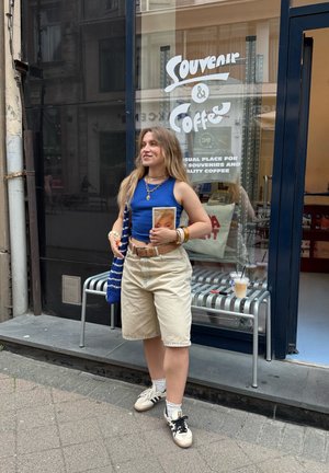 Pantalones cortos beige, top de tirantes azul, cinturón marrón. Los accesorios incluyen collares en capas y pulseras. Sostiene una revista. De pie frente a un café.