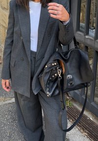 Nő szürke kockás öltönyben és fehér ingben, fekete bőr kézitáskát tart, melyen fém szegecsek vannak, és ezüst gyűrűket visel.