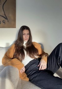 Mustársárga kötött kardigán fehér felsővel, sötét, bő szabású nadrággal párosítva. A modell egy világos színű kanapén ül. Lágy megvilágítás.