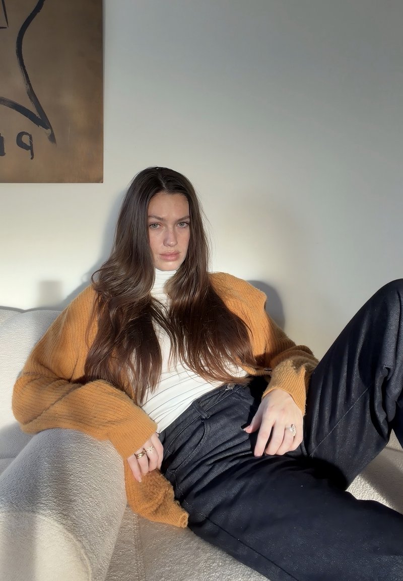 Cardigan lavorato a maglia color senape sopra una maglietta bianca, abbinato a pantaloni scuri e ampi. Modello seduto su un divano chiaro. Illuminazione soffusa.