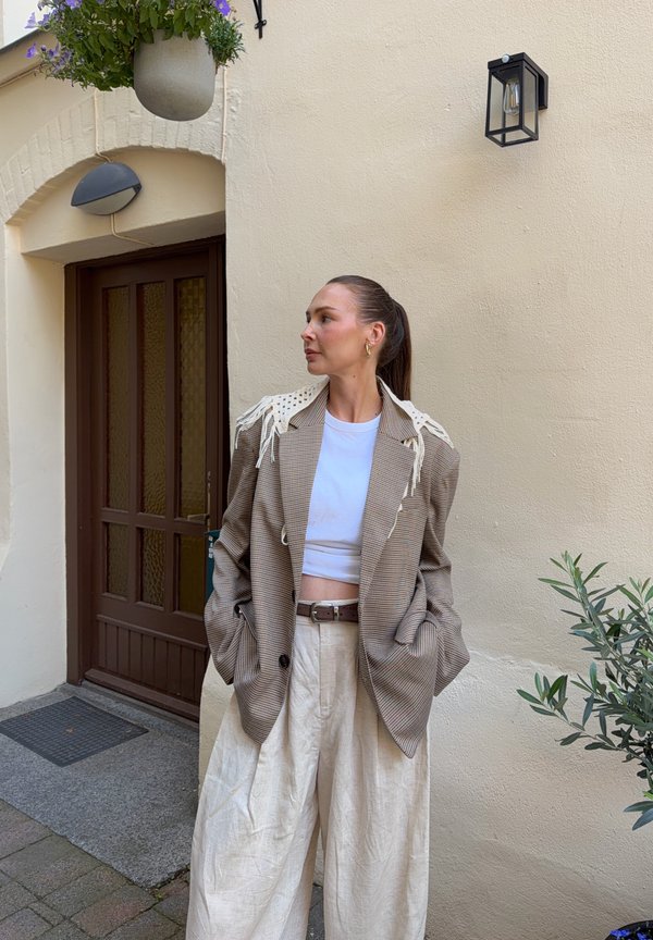 Una mujer con una americana de rayas y pantalones blancos anchos está junto a una puerta marrón, mirando hacia un lado al lado de una pared de estuco color crema.