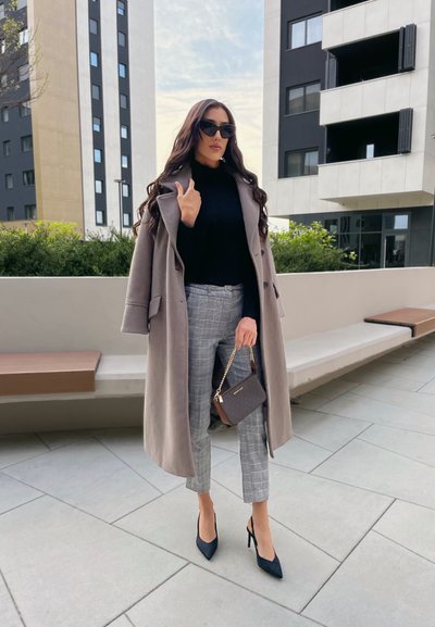 Abrigo de lana gris claro sobre un suéter negro de cuello alto, combinado con pantalones de cuadros grises y tacones negros. Sosteniendo un pequeño bolso oscuro.