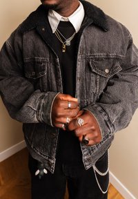 Personne portant une veste en denim noire doublée en shearling, bagues aux doigts, colliers superposés et pantalon noir avec une chaîne argentée et un porte-clés attachés.