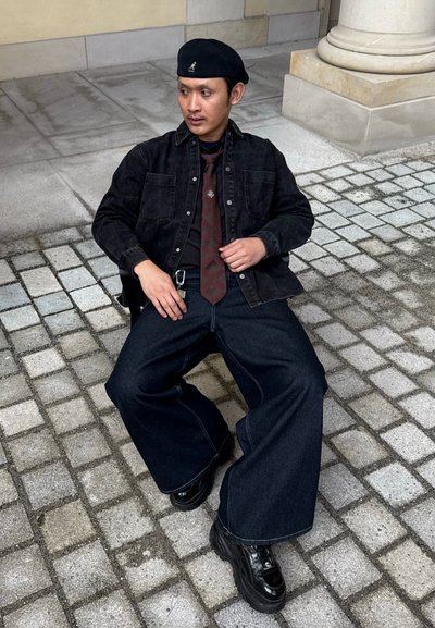 Camisa de mezclilla negra, corbata de patrón burdeos, jeans de pierna ancha negros y zapatos negros gruesos. La persona lleva un boina negra. Entorno exterior texturizado.