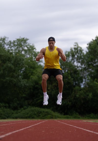 Atleta con una camiseta sin mangas amarilla y pantalones cortos negros salta sobre una pista roja. Lleva zapatillas deportivas blancas con detalles negros y una gorra negra.