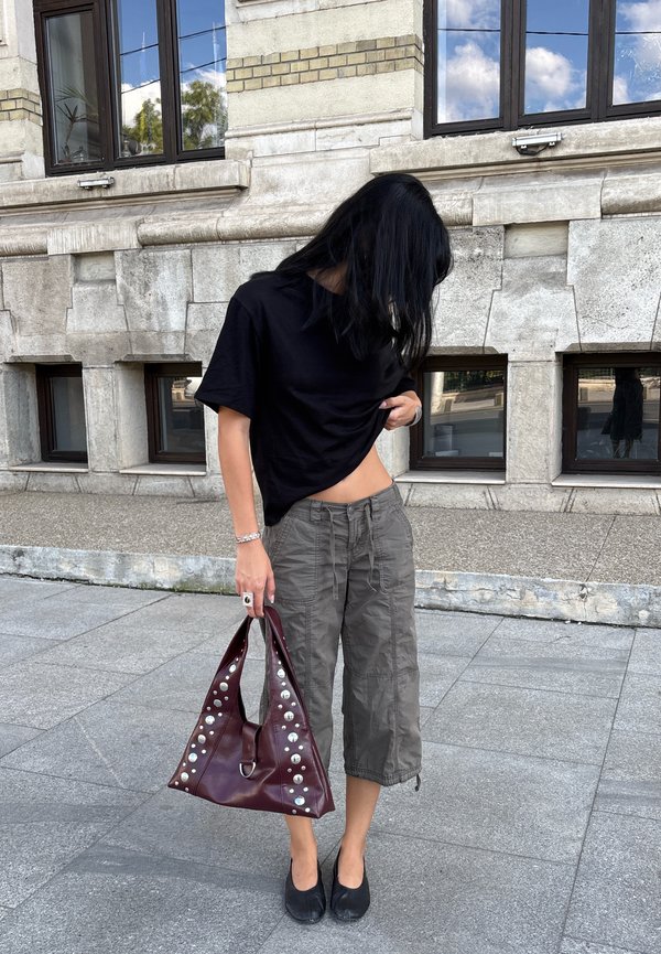 Persona con cabello largo y negro que lleva una camiseta negra y pantalones grises recortados, sostiene un gran bolso burdeos con tachuelas y está de pie sobre un pavimento de piedra.
