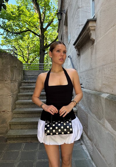 Top negro sin mangas combinado con una falda blanca plisada. La modelo sostiene un pequeño bolso negro con lunares. Escaleras de piedra natural y vegetación de fondo.