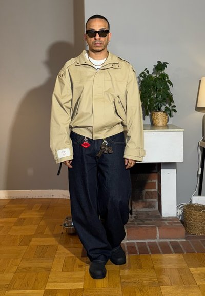 Hombre con chaqueta beige, pantalones anchos oscuros con llaveros de labios y juguetes, zapatos negros, gafas de sol, de pie en el interior junto a una planta y una lámpara.