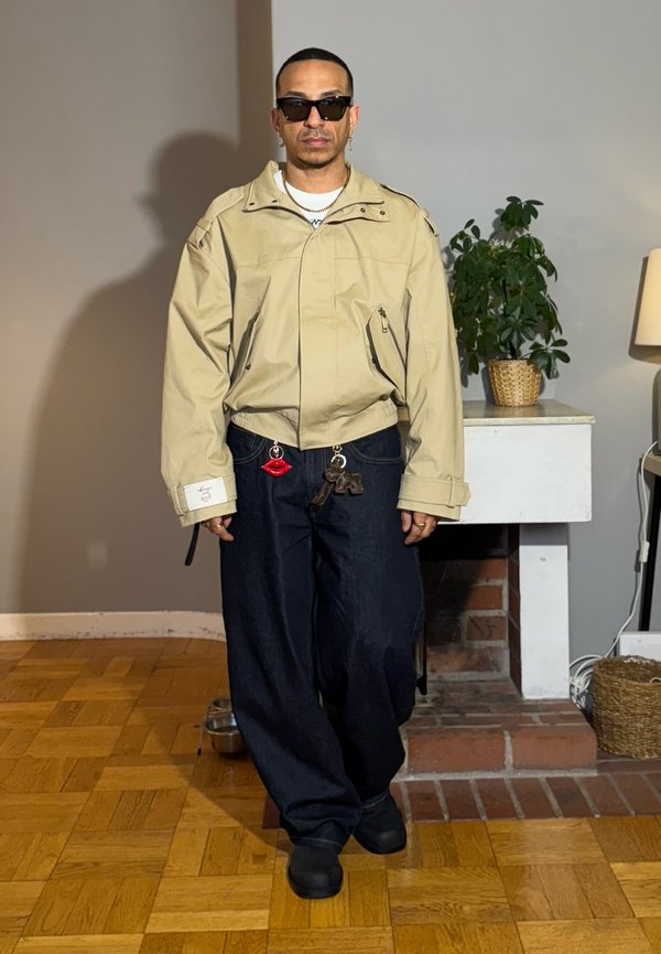 Mand iført beige jakke, mørke vide bukser med læbe- og legetøjs-nøgleringe, sorte sko, solbriller, står indendørs ved en plante og lampe.