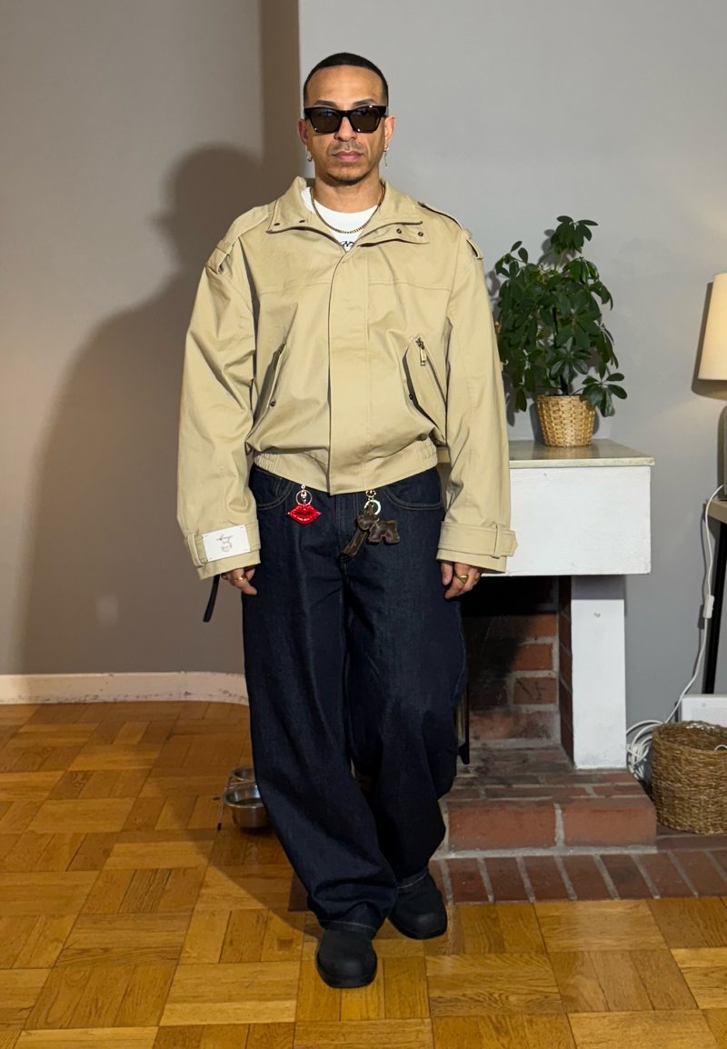 Man wearing beige jacket, dark wide pants with lip and toy keychains, black shoes, sunglasses, standing indoors by plant and lamp.