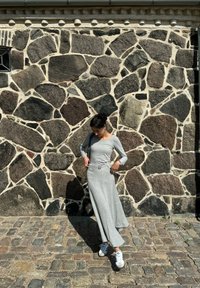 Robe longue grise à manches longues avec une taille ceinturée, fabriquée dans un tissu léger, associée à des baskets blanches. Mur en pierre en arrière-plan avec des textures variées.