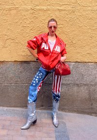 Rdeča jakna z logotipom "PUMA", bela majica, modri kavbojci z ameriškim zastavnim potiskom, srebrni kavbojski škornji z cvetličnim vzorcem, rdeča torbica.