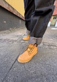 Brune lær arbeidsstøvler med svarte detaljer og gule snørebånd. Støvlene har en solid såle og er kombinert med oppbrettede mørk denim-bukser.