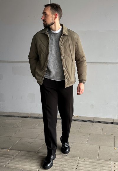 Chaqueta tipo abrigo en verde olivo, suéter de canalé gris debajo, pantalones negros y zapatos negros. Tejido suave, corte relajado y cierre de botones.