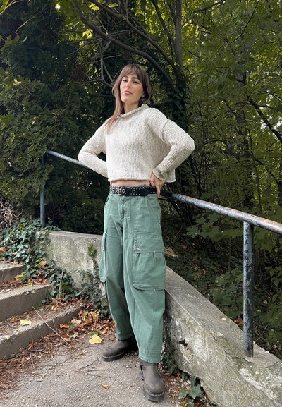 Suéter de punto grueso color crema, pantalones cargo verdes con bolsillos, cinturón negro con tachuelas y botas marrones, todo en un fondo natural.