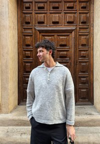 Pulover tricotat gri deschis cu guler cu nasturi, purtat peste o cămașă albă. Fundalul prezintă o ușă texturată din lemn cu panouri ridicate.