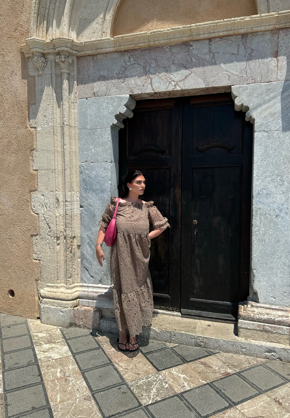 Μακρύ, καφέ λευκό φόρεμα με φουσκωτά μανίκια και στρωμνιστό τελείωμα, σε συνδυασμό με μια ροζ τσάντα. Σκοτεινές ξύλινες πόρτες και τοίχος από υφασμένο πέτρα στο φόντο.