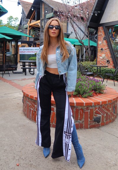 Chaqueta de mezclilla corta, top corto blanco, pantalones deportivos negros con botones de lado y rayas blancas, botas azules de punta, gafas de sol. Entorno al aire libre.