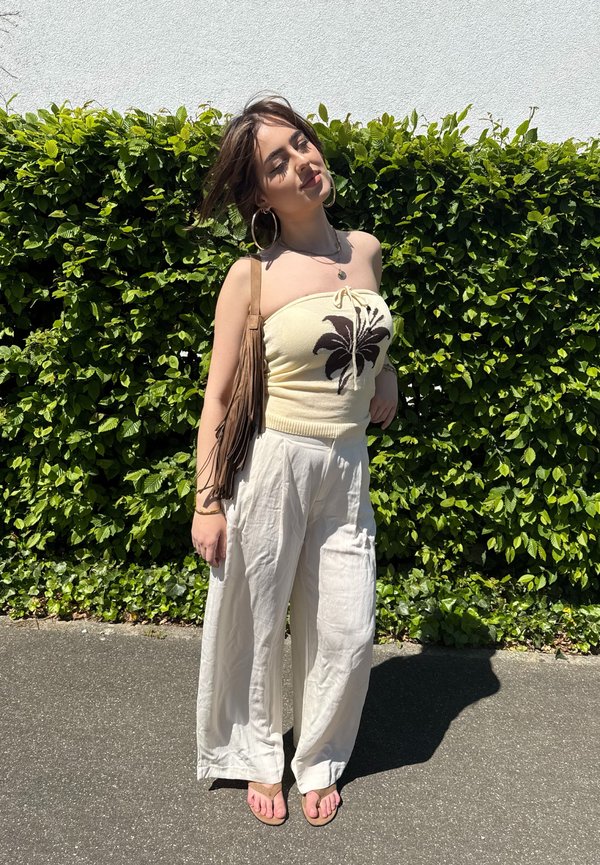 Jeune femme portant un top tube floral crème, un pantalon blanc à jambes larges, des boucles d’oreilles créoles et un sac à franges à l’épaule, debout sur le trottoir à côté d’une haie verte au soleil.