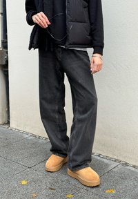 Zwarte puffer vest over een zwarte hoodie, gecombineerd met zwarte losse jeans en tan instapschoenen. Betonnen achtergrond met verstrooide bladeren.