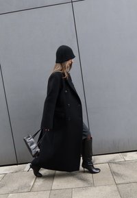 Femme en manteau noir, chapeau noir et bottes à talons noires marchant sur un trottoir carrelé, portant un petit sac à main noir, devant un mur gris.
