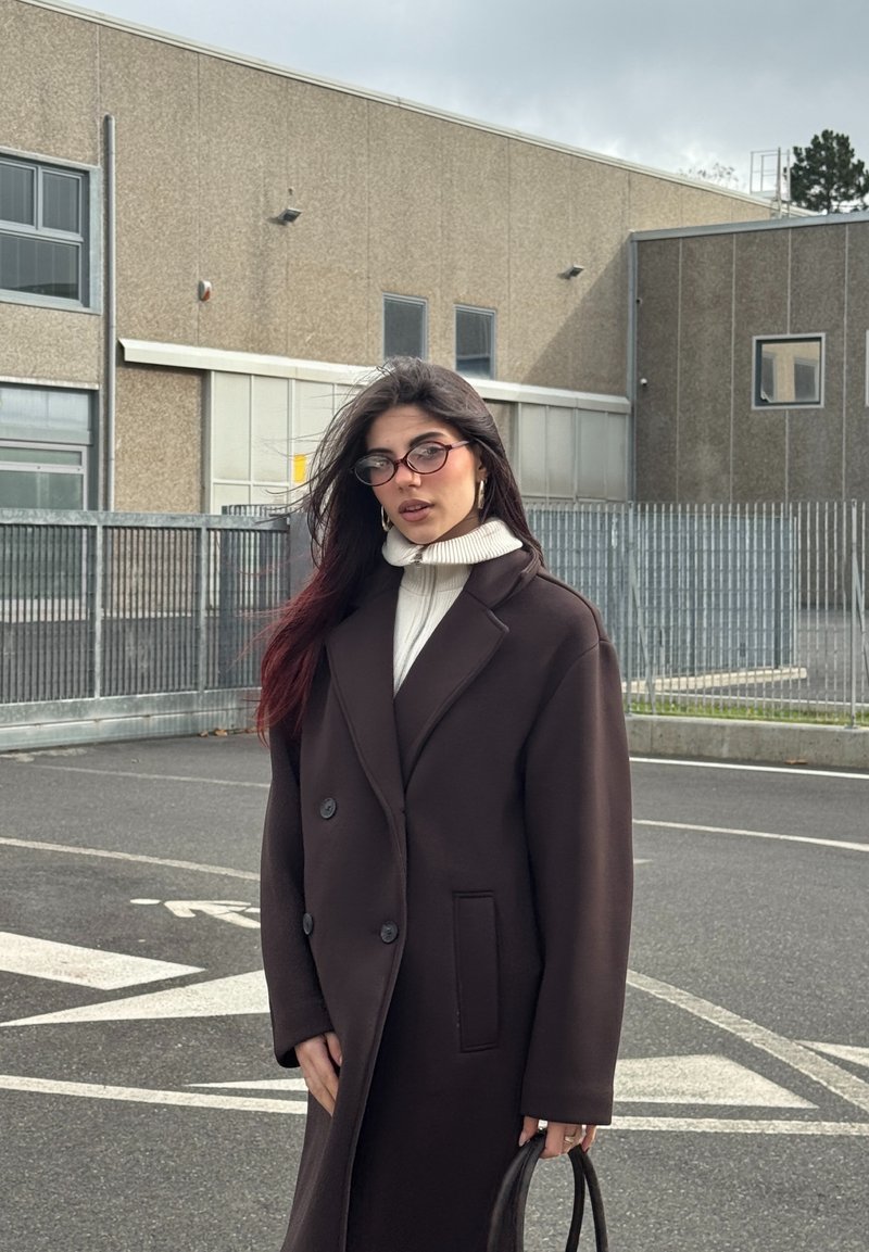 Manteau long marron foncé avec de gros boutons, porté par-dessus un col roulé blanc. Les accessoires comprennent des lunettes roses et un sac à main noir.