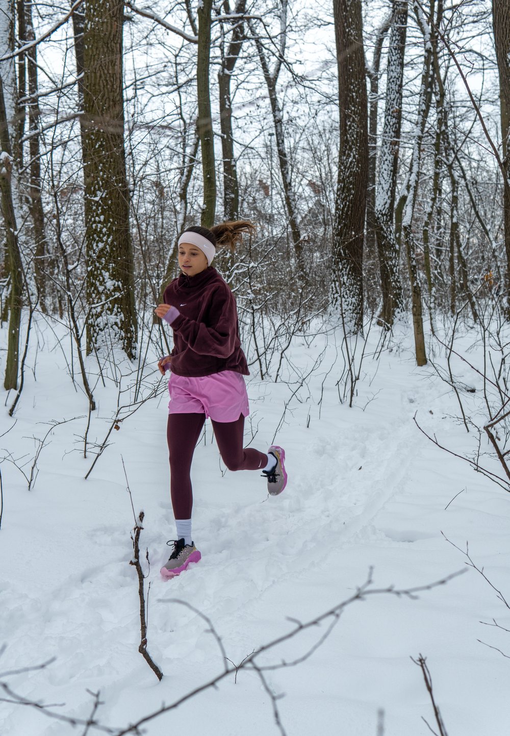 Une femme court à travers une forêt enneigée, vêtue d'une polaire violet foncé, d'un short rose et de leggings bordeaux, avec des chaussures de course roses et noires.