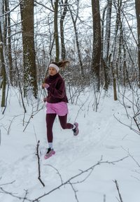 Une femme court à travers une forêt enneigée, vêtue d'une polaire violet foncé, d'un short rose et de leggings bordeaux, avec des chaussures de course roses et noires.