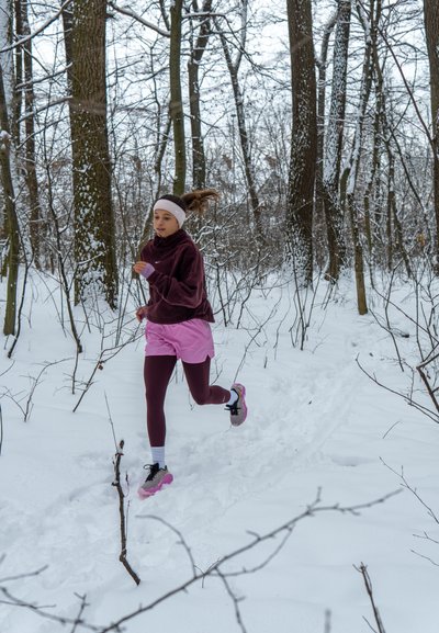 Una mujer corre por un bosque nevado vistiendo un forro polar de color púrpura oscuro, pantalones cortos rosas y leggings granate, con zapatillas de correr rosas y negras.