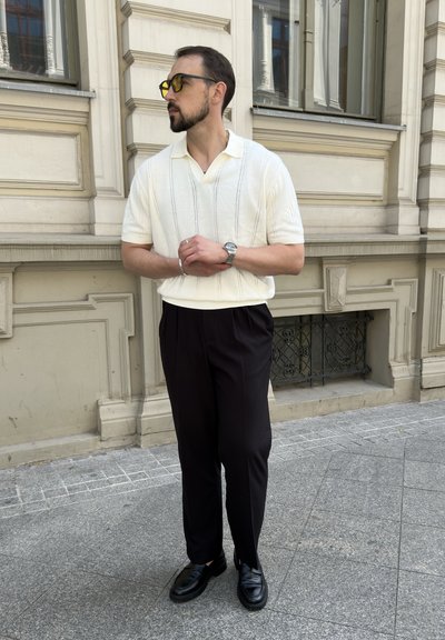 Polo de punto blanco con detalles de rayas verticales, pantalones de vestir negros y zapatos negros. Los accesorios incluyen un reloj y unas gafas de sol con lentes amarillos.