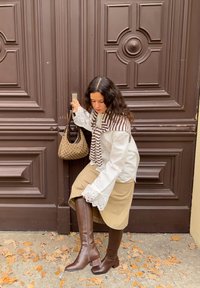 Hvit bluse med stripete brun skjerf, beige skjørt og brune knelange støvler; holder en brun mønstret håndveske ved en brun dør.