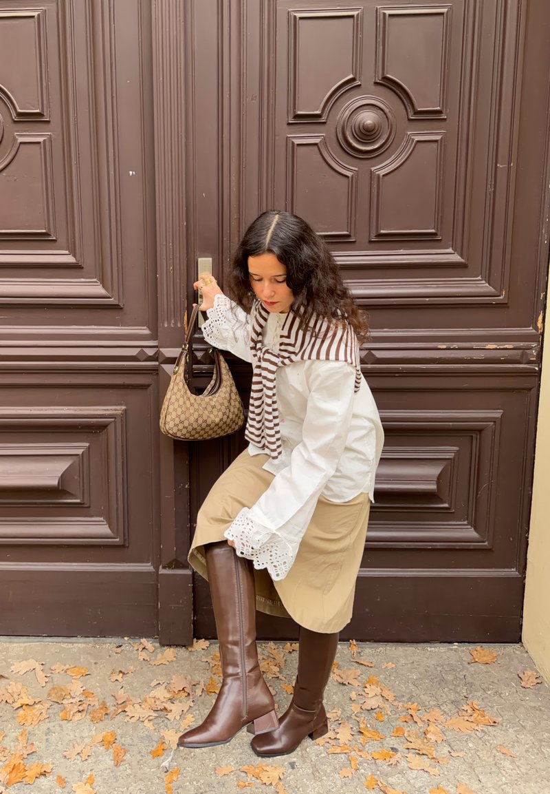 Blusa branca com lenço listrado castanho, saia bege e botas castanhas até ao joelho; segurando uma mala castanha com padrão próxima a uma porta castanha.