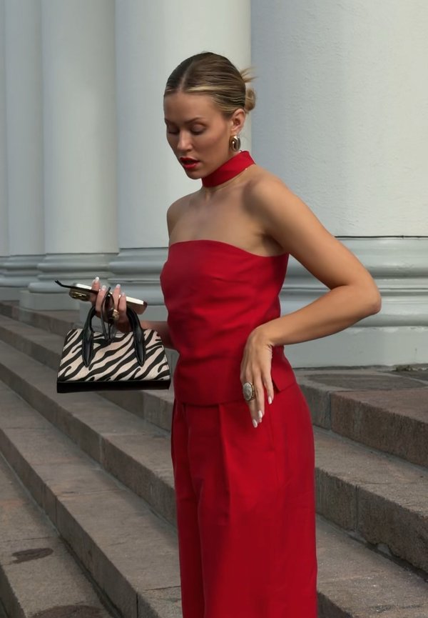 Femme en haut rouge sans bretelles et pantalon tient un sac à main imprimé zèbre et un téléphone, debout sur des marches en pierre près de colonnes blanches.