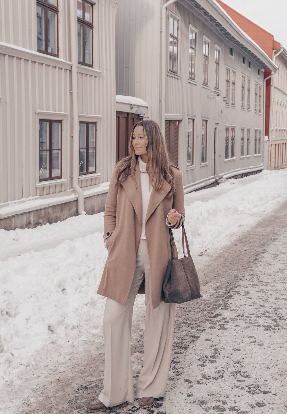Femme en manteau beige et pantalon blanc tenant un sac, debout dans une rue enneigée avec de grands bâtiments en bois clair en arrière-plan.