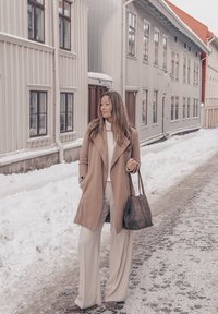Femme en manteau beige et pantalon blanc tenant un sac, debout dans une rue enneigée avec de grands bâtiments en bois clair en arrière-plan.