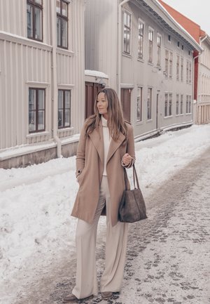 Mujer con abrigo beige y pantalones blancos sosteniendo una bolsa, de pie en una calle nevada con altos edificios de madera clara en el fondo.