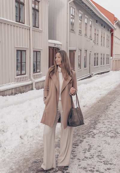 Mujer con abrigo beige y pantalones blancos sosteniendo una bolsa, de pie en una calle nevada con altos edificios de madera clara en el fondo.