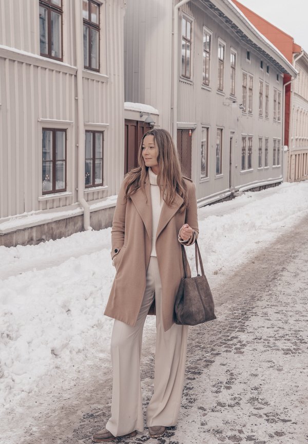 Egy nő bézs színű kabátban és fehér nadrágban, táskát tartva, egy havas utcán áll, háttérben magas, világos színű faépületekkel.