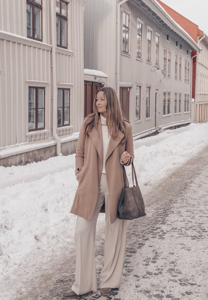 Kvinde i beige frakke og hvide bukser, der holder en taske, stående på en snedækket gade med høje, lyse trækonstruktioner i baggrunden.