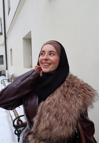 Jeune femme souriante en plein air en hiver, portant un hijab noir, une étole en fausse fourrure marron et une veste en cuir marron foncé près de bancs couverts de neige.