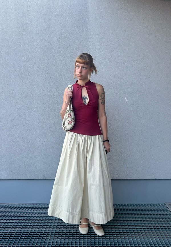 Joven mujer con tatuajes lleva un top burdeos sin mangas y una falda larga blanca, sostiene una bolsa floral, de pie contra una pared gris.