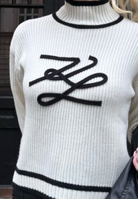 Pull blanc côtelé avec col montant et ourlet bordés de noir, présentant un design abstrait de corde noire sur la poitrine.