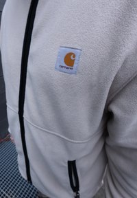 Κρεμ μπουφάν φλις με μαύρο φερμουάρ και τσέπες, με ένα μικρό πορτοκαλί λογότυπο Carhartt στο στήθος.