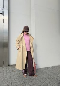 Beige trenchcoat over en rosa genser, brune joggebukser med hvite striper, brun caps og solbriller. Står mot en lys grå vegg.