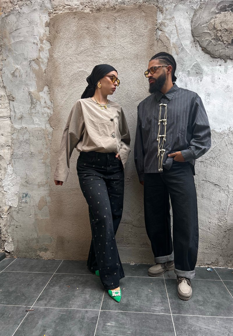 Two people stand against a textured wall, wearing stylish outfits and sunglasses, one in green heels and the other in beige shoes.