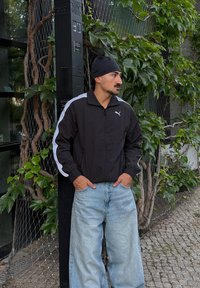 Moški v črni jakni Puma in svetlo modrih širokih hlačah stoji s rokama v žepih blizu žive meje in droga.