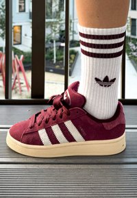 Bordeauxfarbene Wildleder-Sneaker mit weißen Streifen, abgerundetem Zehenbereich und Gummisohle. Getragen mit weißen Socken, die bordeauxfarbene Streifen und ein Logo haben.